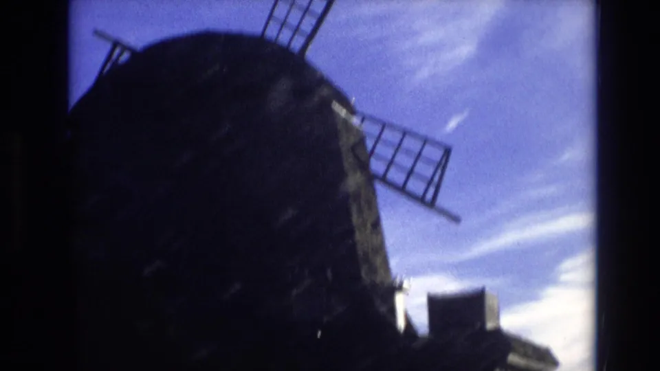Low-angle 1974 Vail, Colorado 8mm view of the windmill dome, blade frame, nearby roofline, and bright clouded sky.