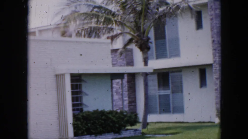 Two-story white brick mid-century modern building in Cuba, c.1956, with palm tree, blue-green louvered windows, and manicured hedges.