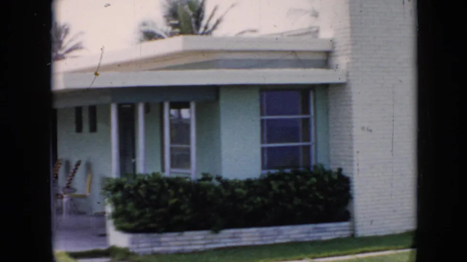 1956 Cuba home-movie frame showing a pastel green bungalow with a covered patio, low hedge, windows, and lounge chairs.