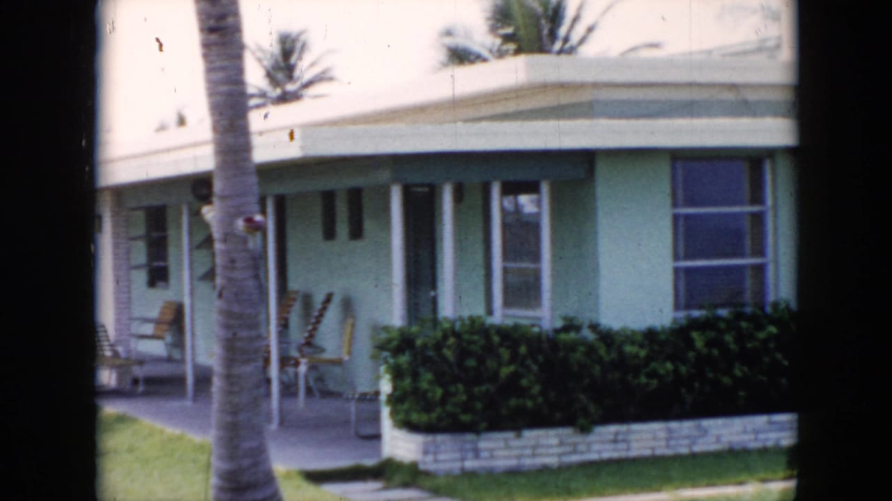 Palm trunk in front of a pastel bungalow with patio chairs, windows, white posts, and hedge.