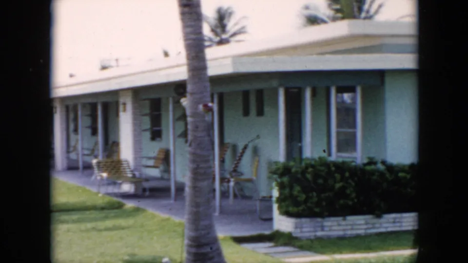 1956 Cuba home-movie frame showing connected bungalow patios, a palm trunk, green lawn, lounge chairs, and low hedging.