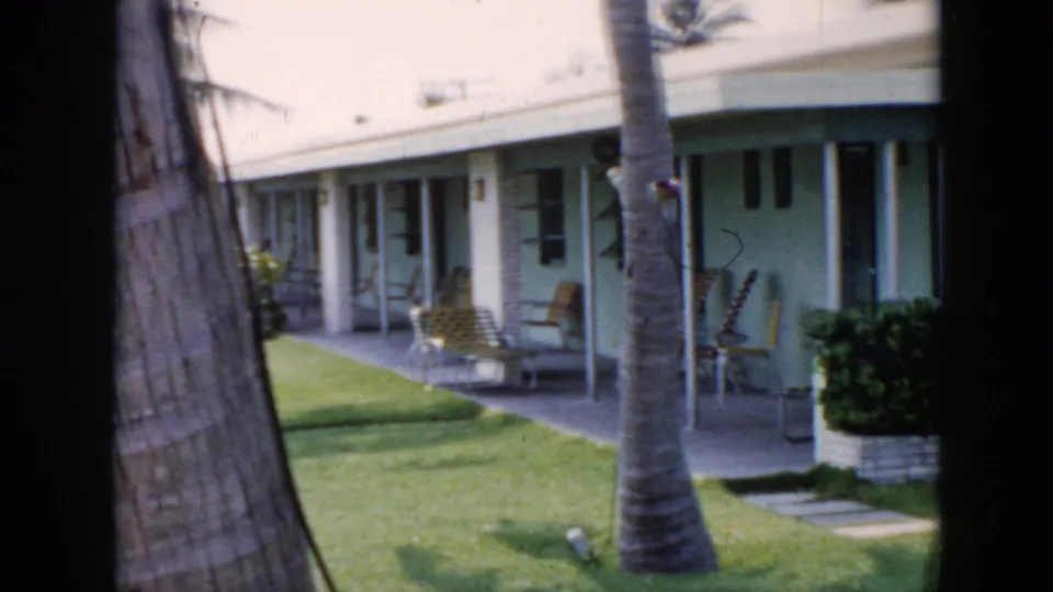 1956 Cuba frame showing an angled view of pastel bungalow patios, palm trunks, lounge chairs, windows, and a sunlit lawn.