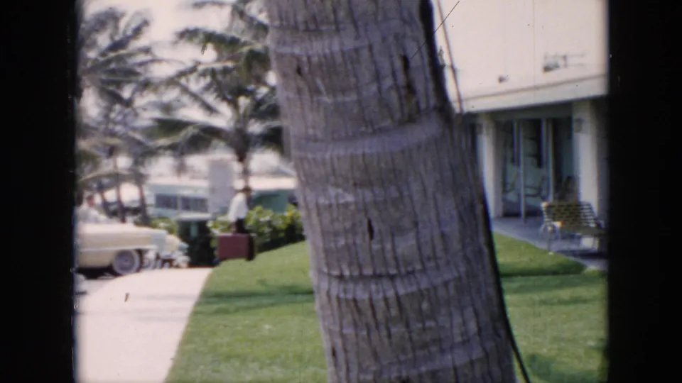 1956 Cuba resort scene with a person near a walkway, palm trees, parked mid-century cars, and motel buildings in the background.