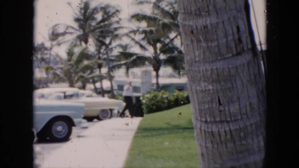 1956 Cuba frame showing classic cars parked near motel grounds, with palms, lawn, walkway, and low resort buildings visible.