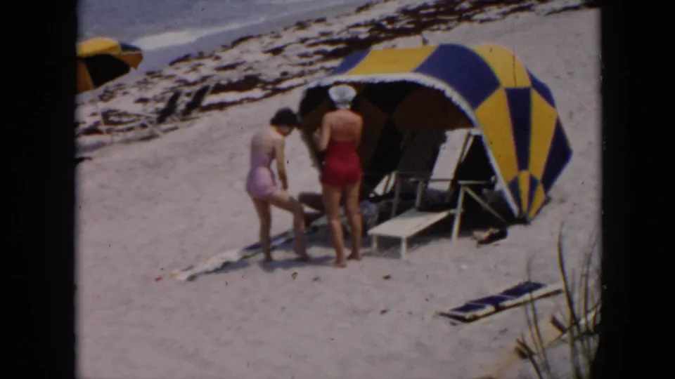 1956 Cuba beach outing frame with two women near a yellow-blue striped tent, beach chairs, sand, and scattered personal items.