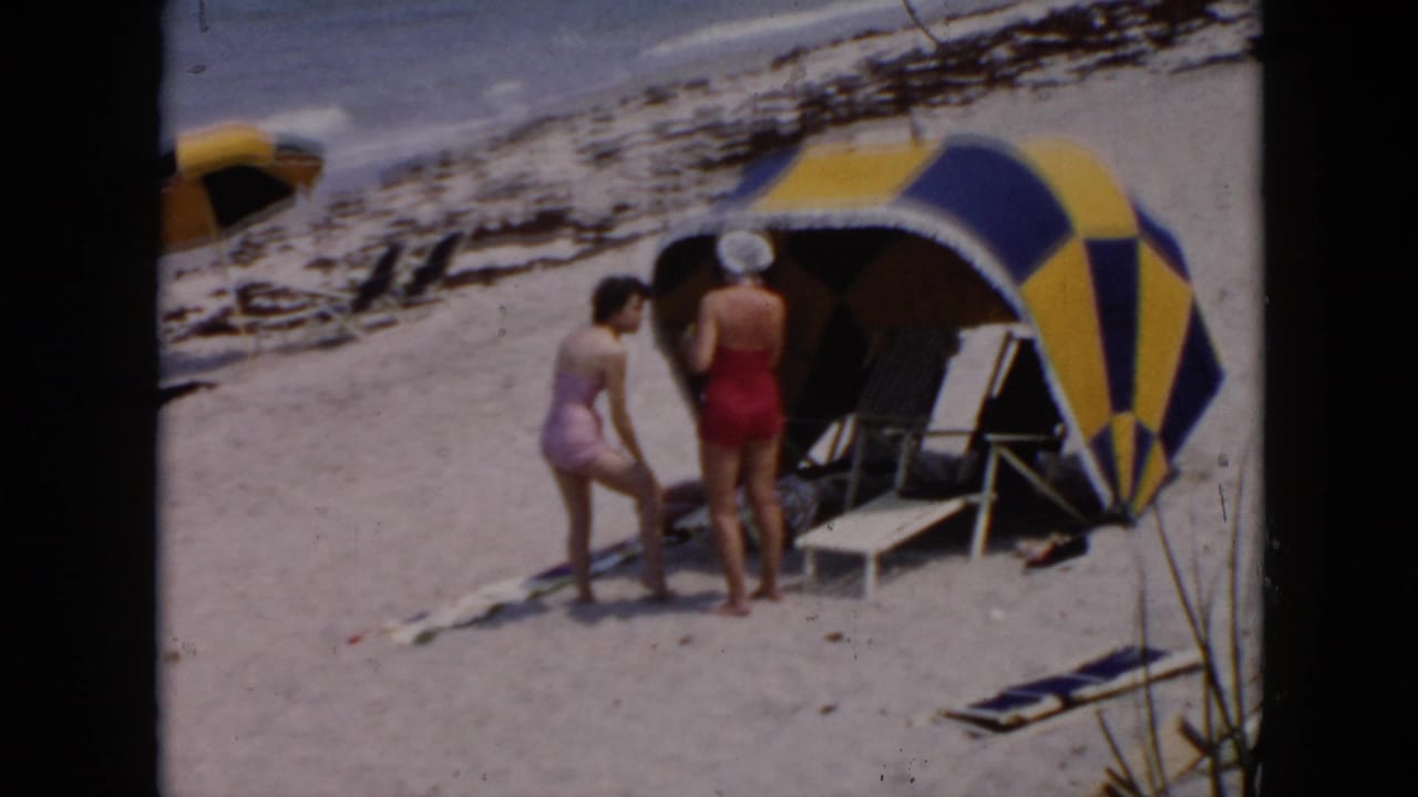 1956 Cuba beach scene with two women in swimsuits near a striped cabana, lounge chairs, and surf in the background.
