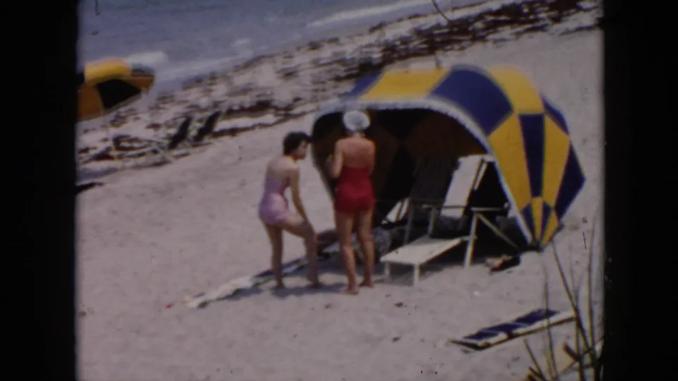 1956 Cuba beach scene with two women in swimsuits near a striped cabana, lounge chairs, and surf in the background.