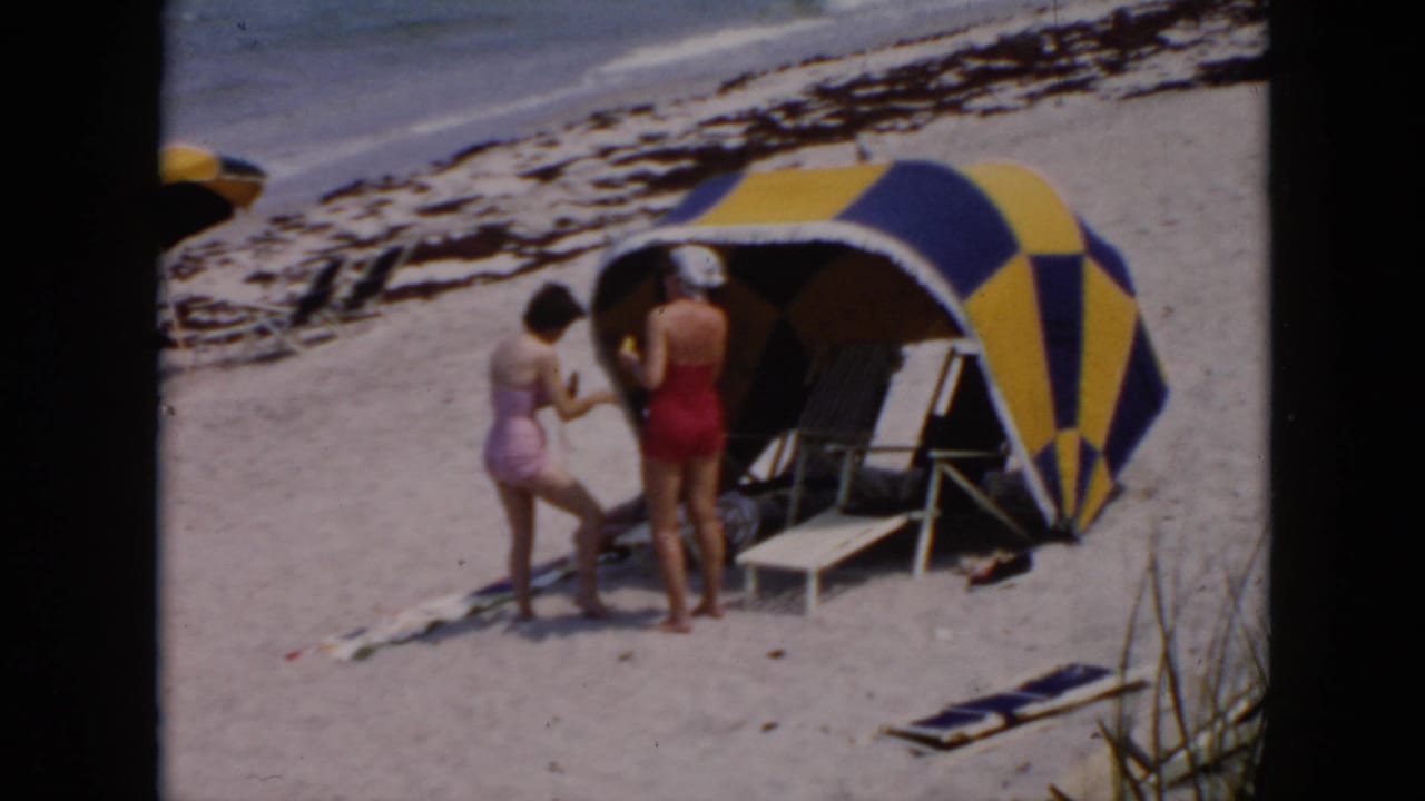 Two women at a Cuba beach cabana, one in lavender leaning toward a woman in red near folding chairs and surf.