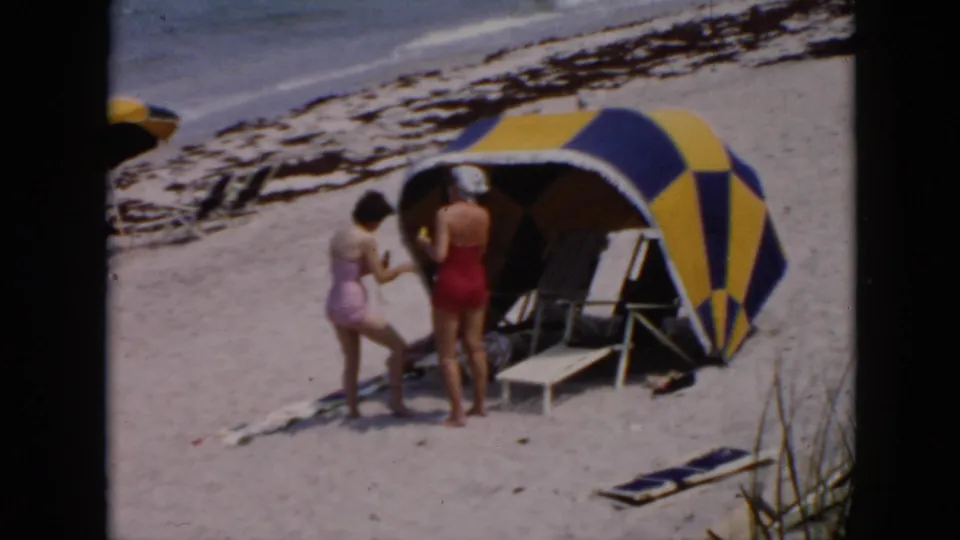 Two women at a Cuba beach cabana, one in lavender leaning toward a woman in red near folding chairs and surf.