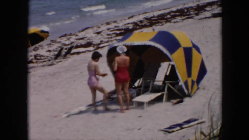 1950s home-movie frame of two women beside a blue and yellow beach cabana with chairs, sand, and surf visible.