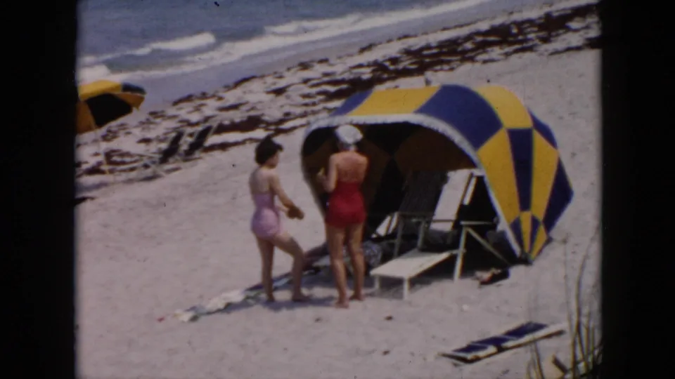Cuba beach home-movie frame with two women near a striped cabana, folding chairs, beach mats, dune grass, and surf.
