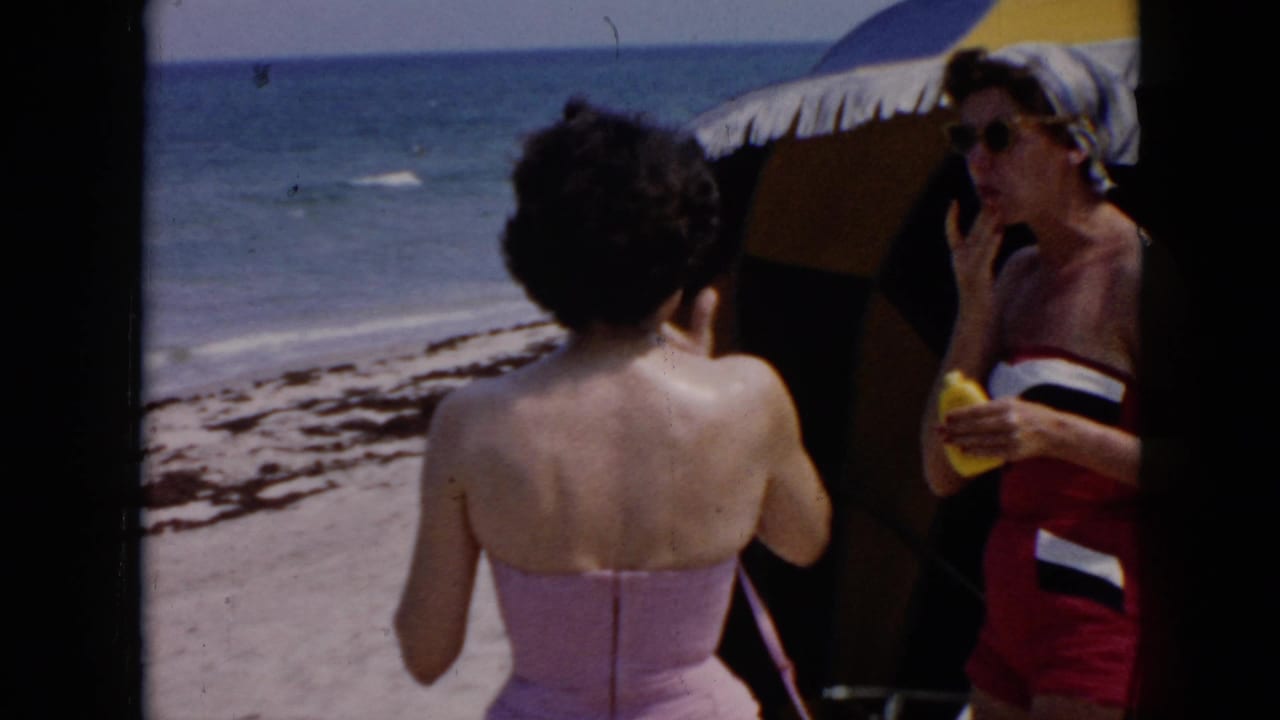 Cuba 1956 home-movie frame showing two adult women on a beach, one turned away in a pink swimsuit and another near an umbrella with sunscreen.
