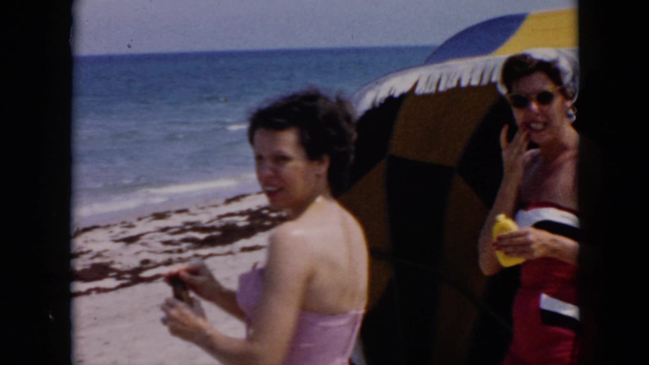 A woman in a pink swimsuit turns toward the camera near another woman under a beach umbrella.