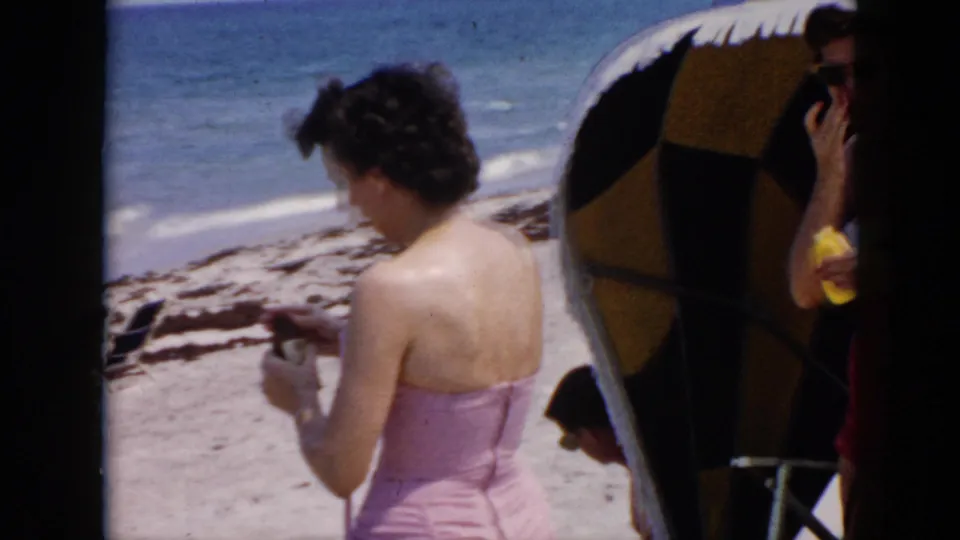 Cuba 1956 home-movie beach frame showing a woman in a pink swimsuit looking down near sand and surf, with another woman partly hidden by an umbrella.