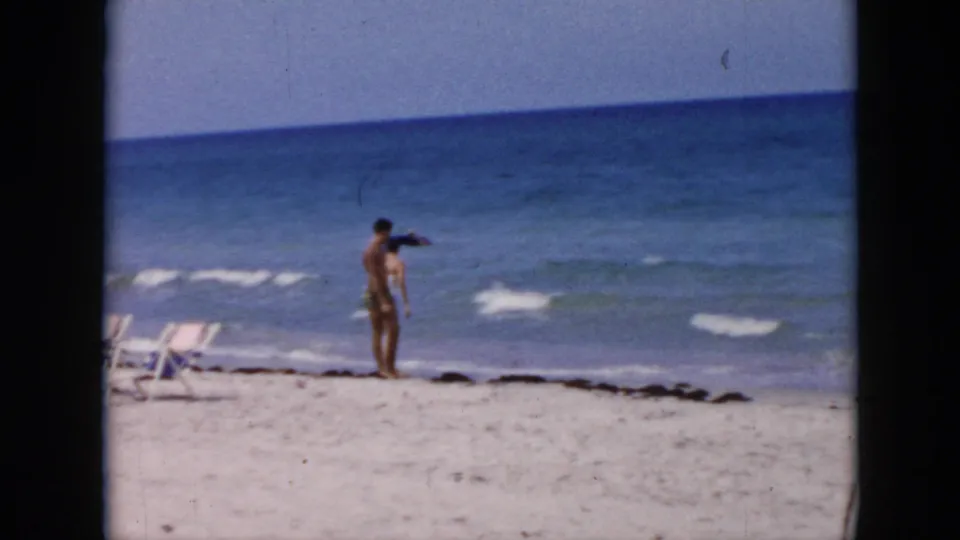 Cuba 1956 beach frame showing an adult man near the surf, with empty lounge chairs, sand, seaweed, and blue water in the background.
