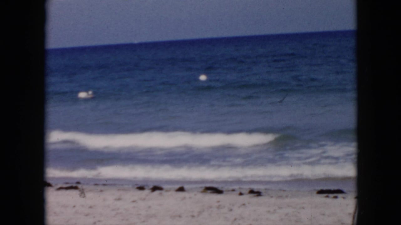 1956 Cuba 8mm frame showing blue ocean, gentle surf, sandy shoreline, and a small white buoy in the distance.