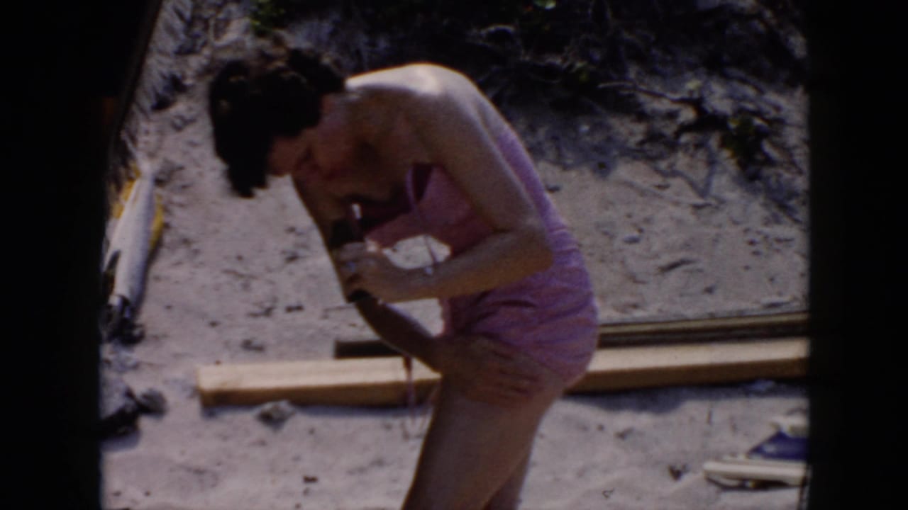 Woman in a pink swimsuit bends over on a sandy beach while applying sunscreen.