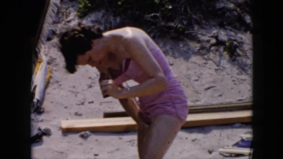 1956 Cuba beach home-movie frame showing the woman in a pink swimsuit bent forward while applying sunscreen beside wooden planks and sand.