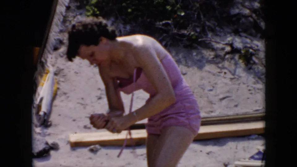 1956 Cuba 8mm frame of the woman rubbing sunscreen onto her leg, with wood planks, sand, and beach debris visible around her.