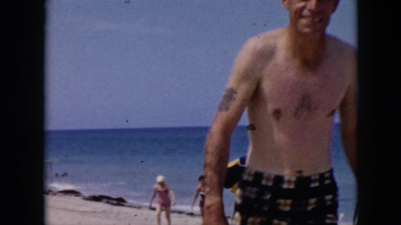 In supported 1956 Cuba home-movie footage, a shirtless man in plaid swim trunks walks close to the camera on a sunny beach with swimmers in the distance.