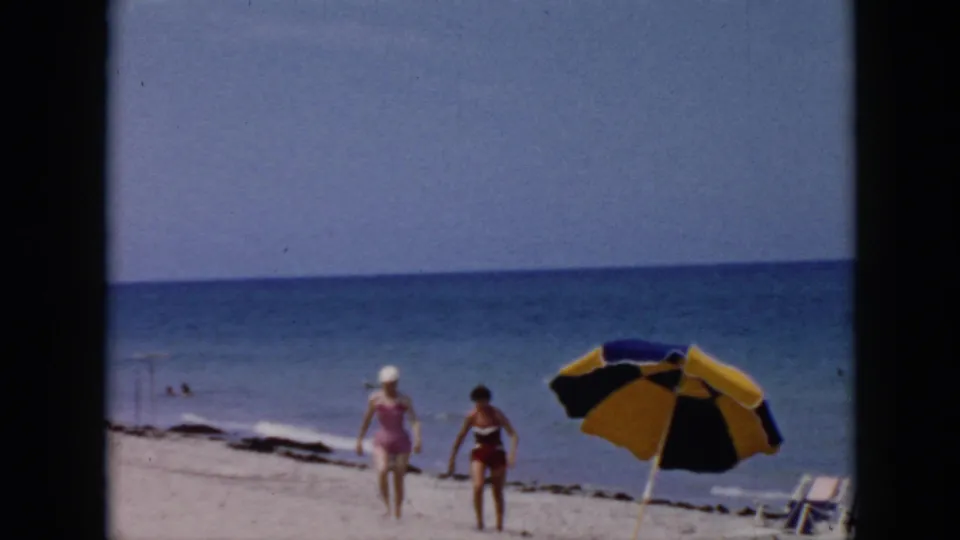 Two women in retro swimsuits walk along a Cuba beach, with a yellow and dark umbrella, lounge chairs, sand, and blue ocean visible behind them.