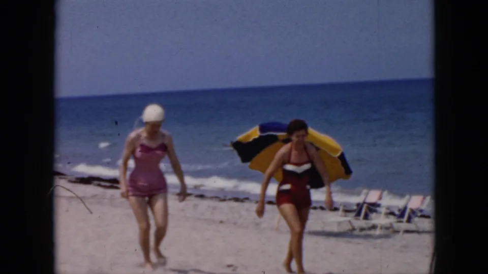 A woman in a pink one-piece and white swim cap walks beside a woman in a red and white two-piece on a sunny Cuba beach.