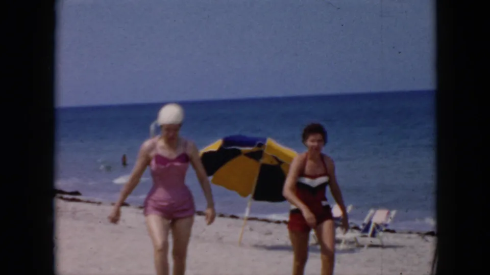 Two adult women in mid-century swimsuits walk along the sand in supported 1956 Cuba footage, framed by a blue ocean horizon and beach furniture.
