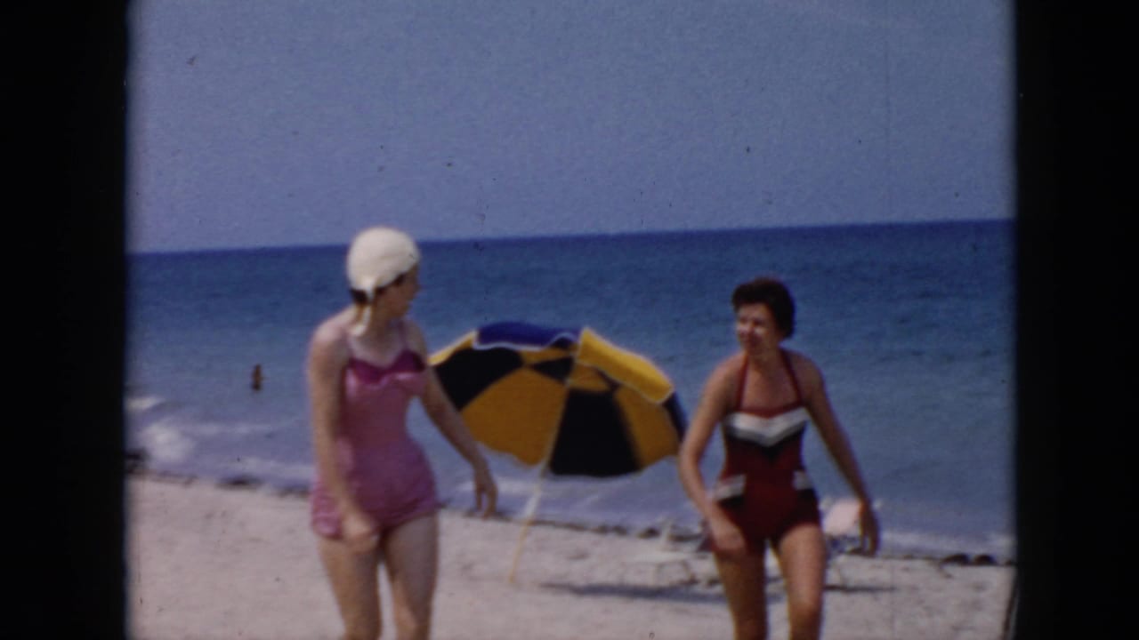 Two women in 1956 Cuba beach footage walk along sand near a yellow and black umbrella, with waves behind them.