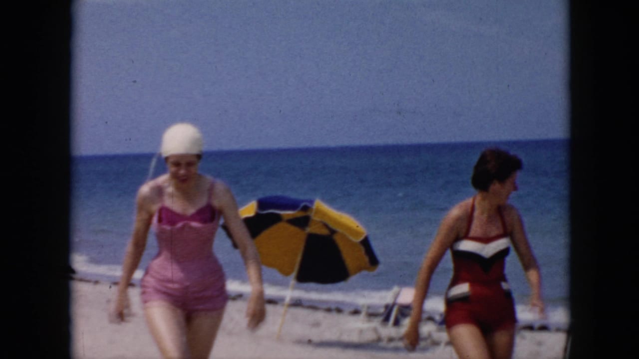 Two women walk separately across a sandy beach with ocean and umbrella behind them.