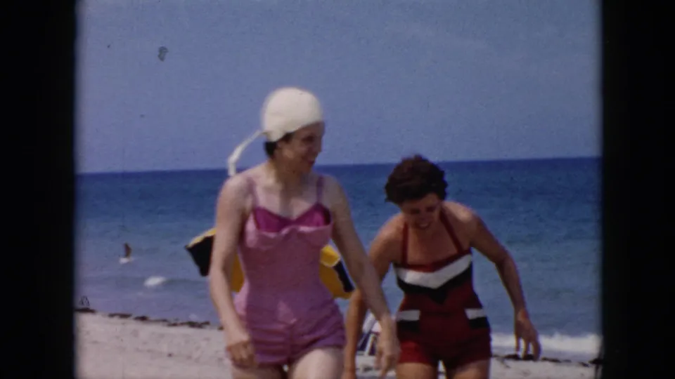 Two women in 1956 Cuba home-movie beach footage approach the camera, with ocean, sand, and umbrella visible.