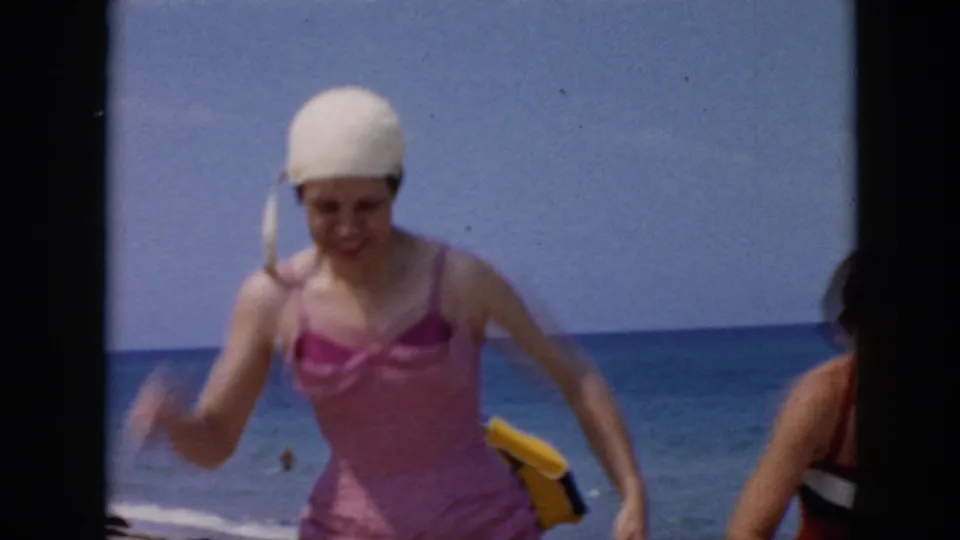 A woman in a pink one-piece swimsuit and white swim cap passes close to the camera in 1956 Cuba beach footage.