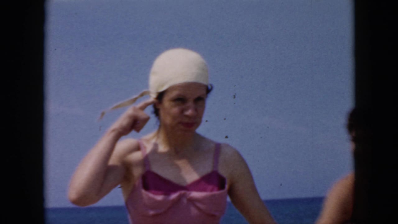 1956 Cuba home-movie frame showing a woman in a pink swimsuit adjusting a white swim cap with blue sky and sea behind her.