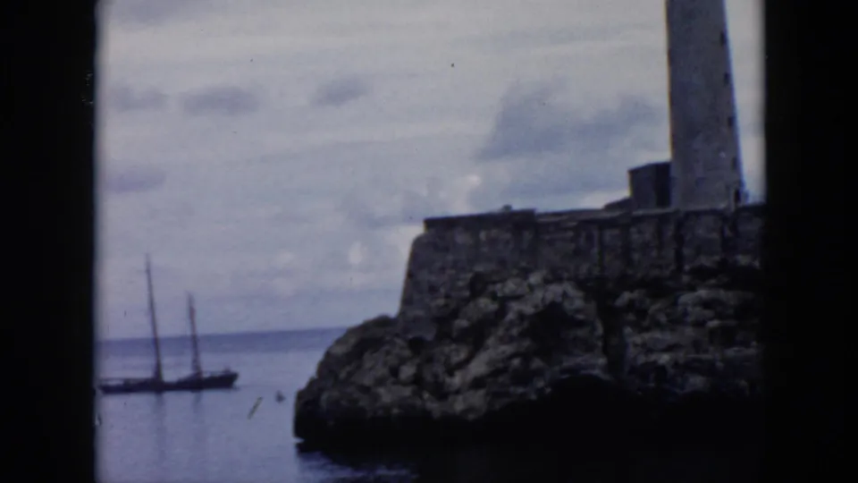 1956 Cuba home-movie view of a stone coastal fortification with a lighthouse above rocks and a boat on the water.