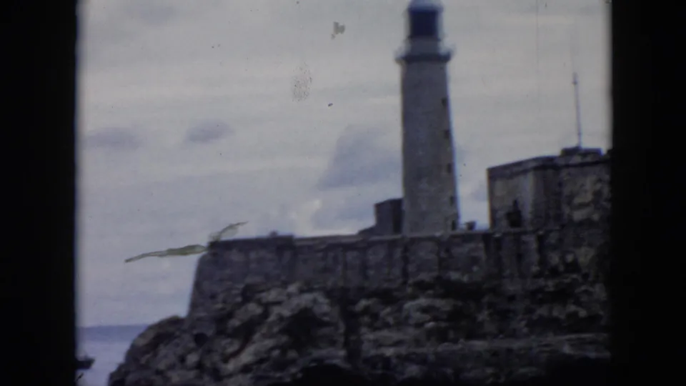 Distant 1956 Cuba coastal frame showing a lighthouse rising from a stone structure on a rocky shore.