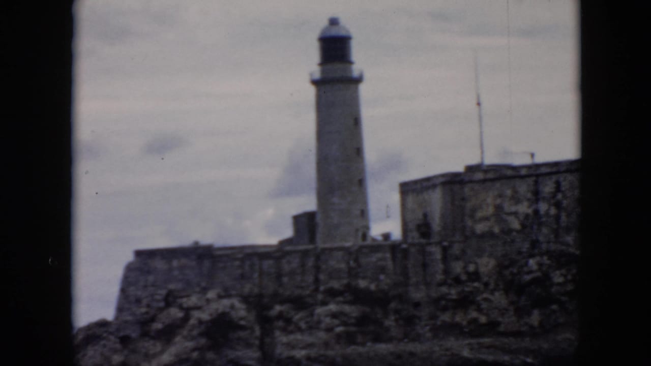 1956 Cuba 8mm frame showing a tall lighthouse behind stone fortress walls under a cloudy sky.
