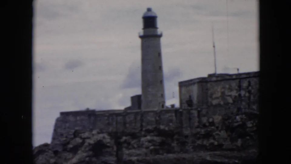 1956 Cuba 8mm frame showing a tall lighthouse behind stone fortress walls under a cloudy sky.