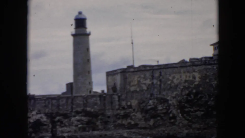 1956 Cuba home-movie frame with the lighthouse centered near fortress masonry and cloudy sky.