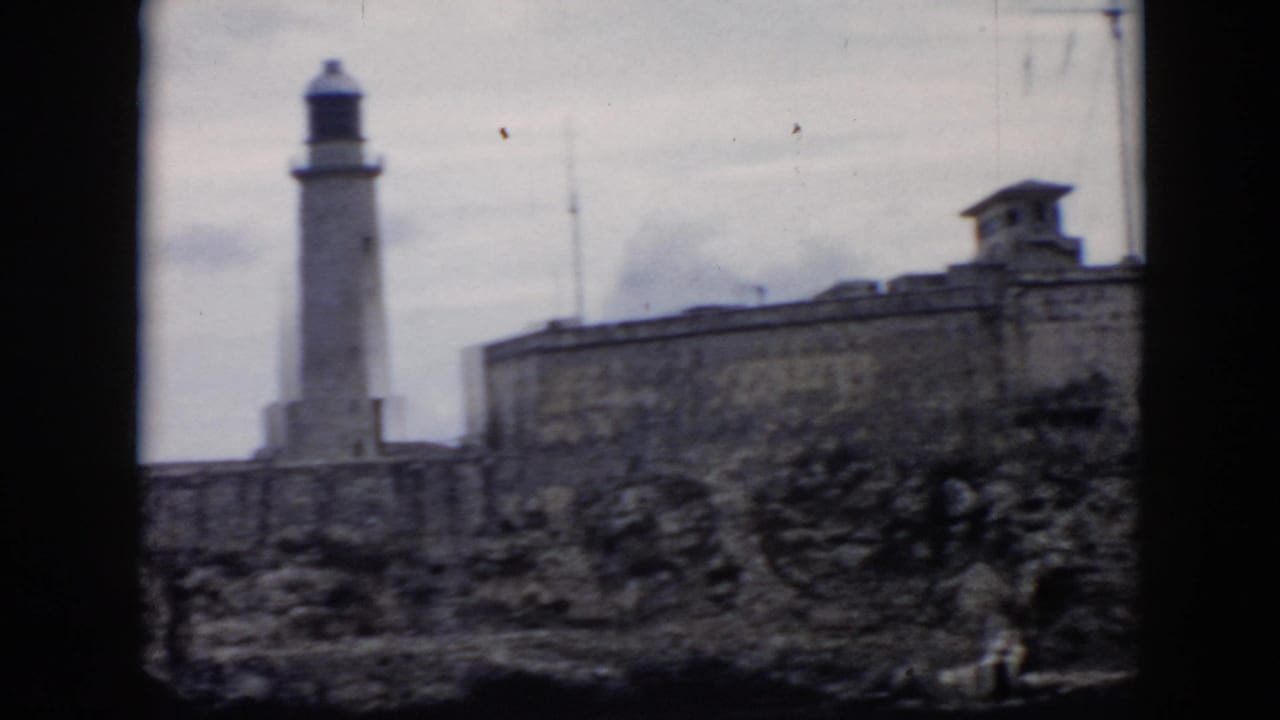1956 Cuba home-movie frame showing a stone fortress wall and lighthouse tower beyond rocky ground under overcast sky.