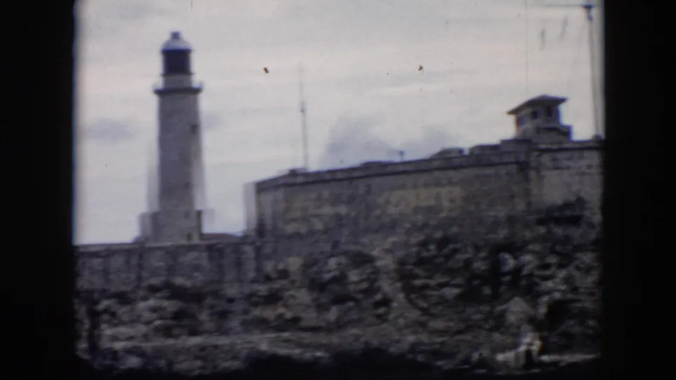1956 Cuba home-movie frame showing a stone fortress wall and lighthouse tower beyond rocky ground under overcast sky.