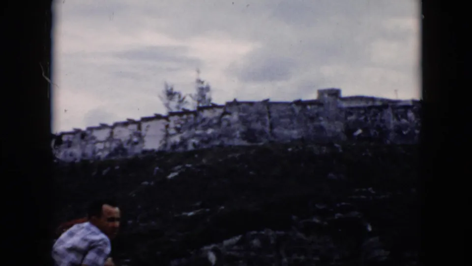 1956 Cuba home-movie frame with a man in a patterned shirt standing below a long stone fortress wall on rocky ground.