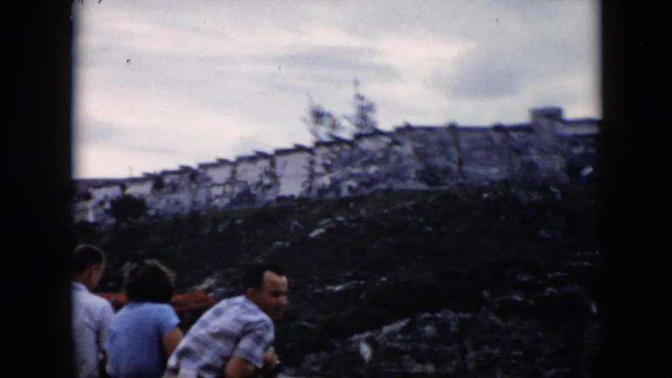 1956 Cuba frame showing tourists gathered on rocky ground below a weathered stone fortress wall under an overcast sky.