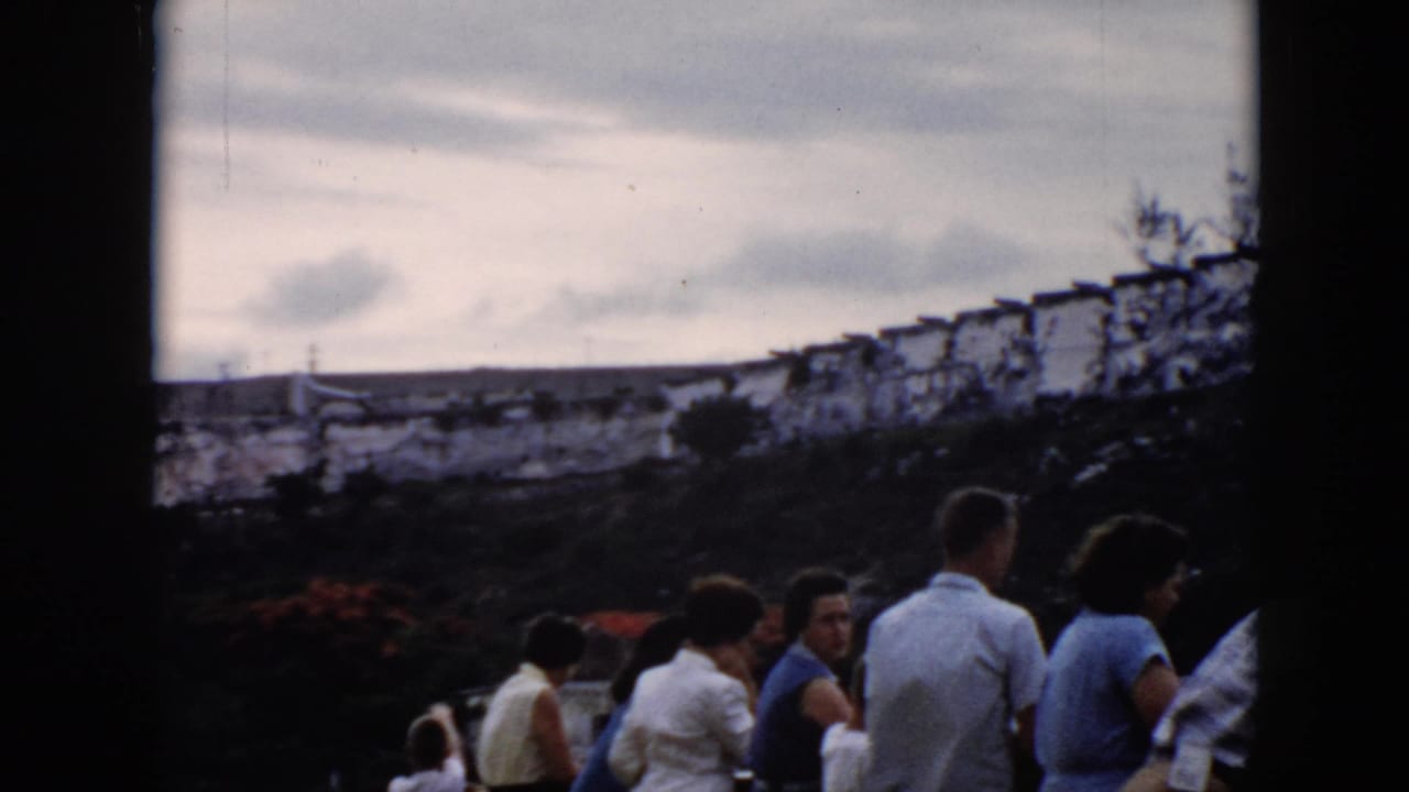 1956 Cuba home-movie frame showing tourists gathered at an overlook with a distant stone fortress and water beyond.