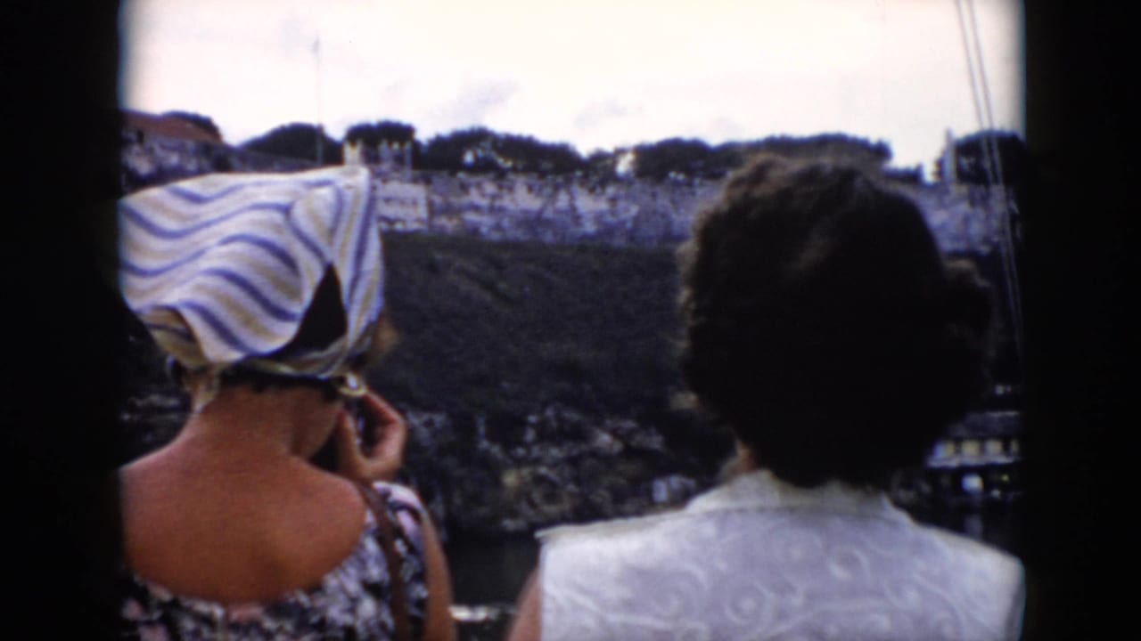 Two women seen from behind look toward a fortress wall, one wearing a striped headscarf.