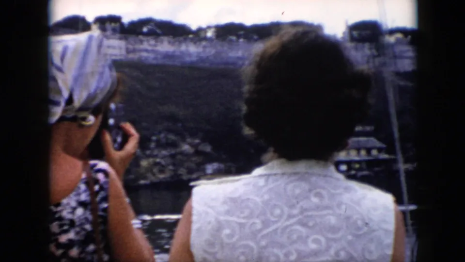 1956 Cuba home-movie frame showing a tourist preparing to take a photo as another woman watches the fortress across the water.