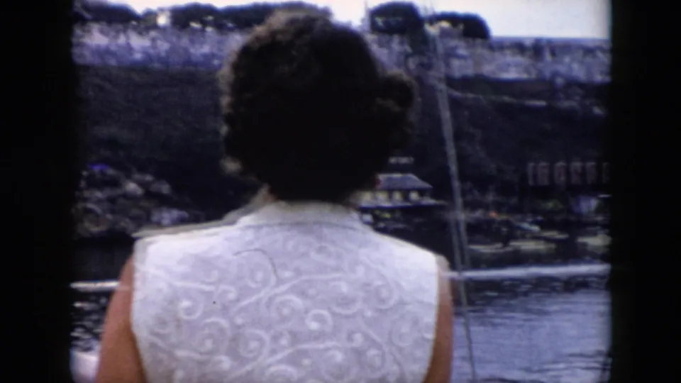 In 1956 Cuba footage, a woman aboard a boat looks toward a riverside wall, buildings, water, and a visible Ironbeer sign.