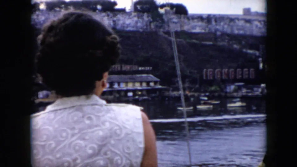 A woman aboard a riverboat in Cuba faces the shoreline, with water, a stone bank, small boats, and an Ironbeer sign visible beyond her.