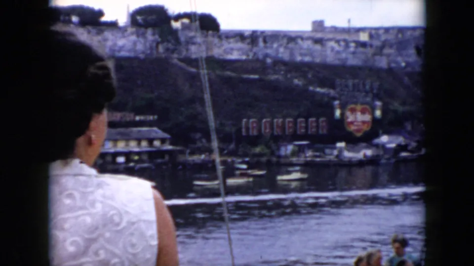 A cropped passenger view from a Cuba riverboat shows water, a waterfront wall, moored boats, and the Ironbeer sign along the shore.