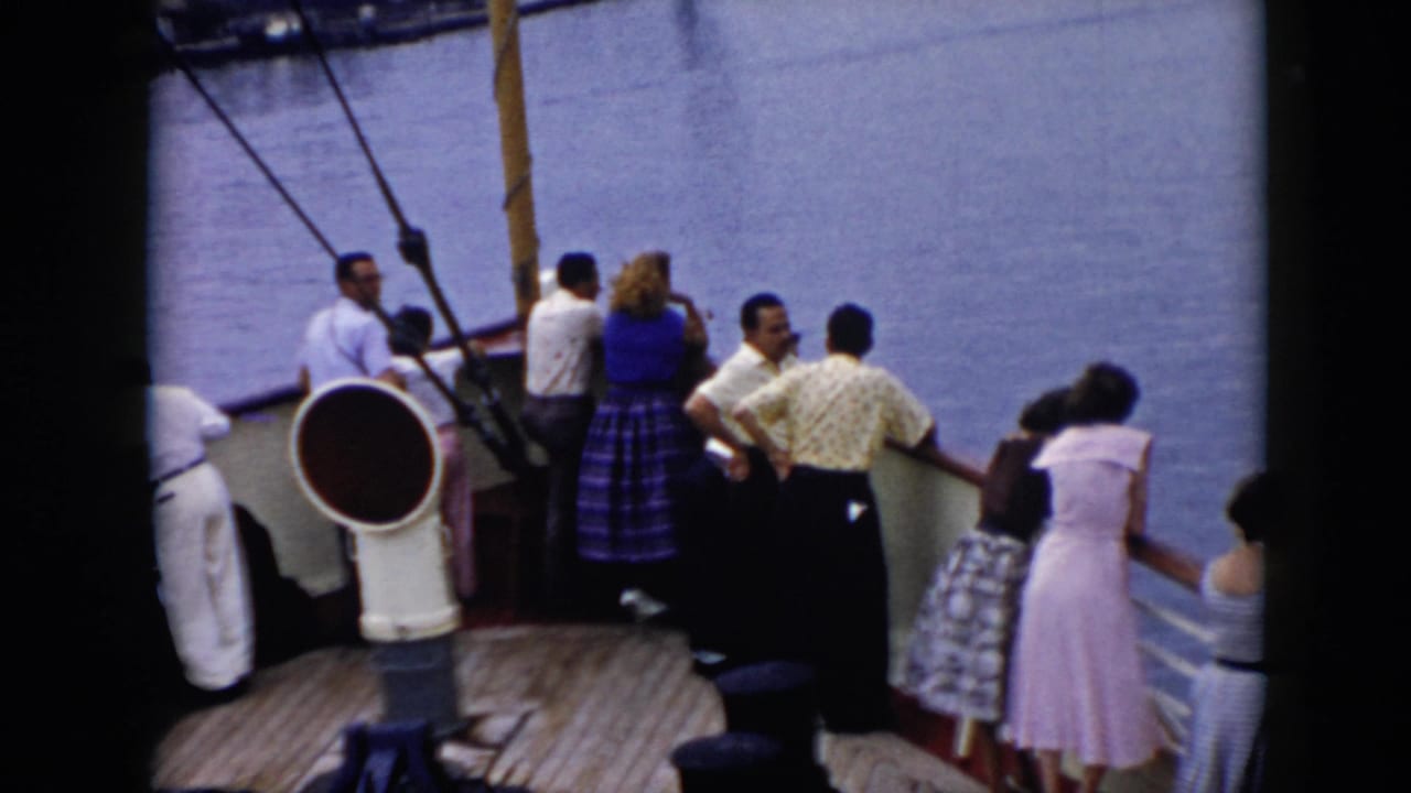 Passengers on a cruise ship deck face the water, with a large white deck fixture and rigging visible in the foreground.