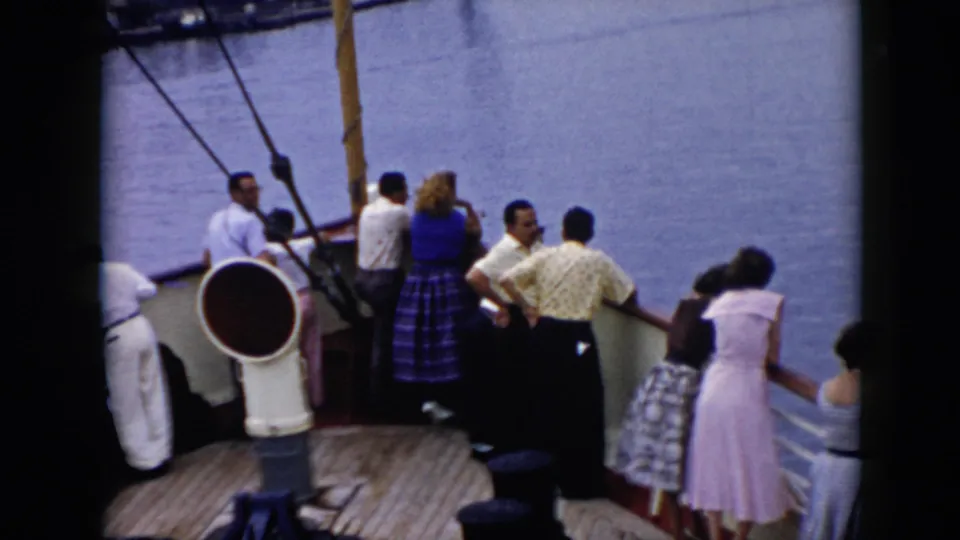 Passengers on a cruise ship deck face the water, with a large white deck fixture and rigging visible in the foreground.