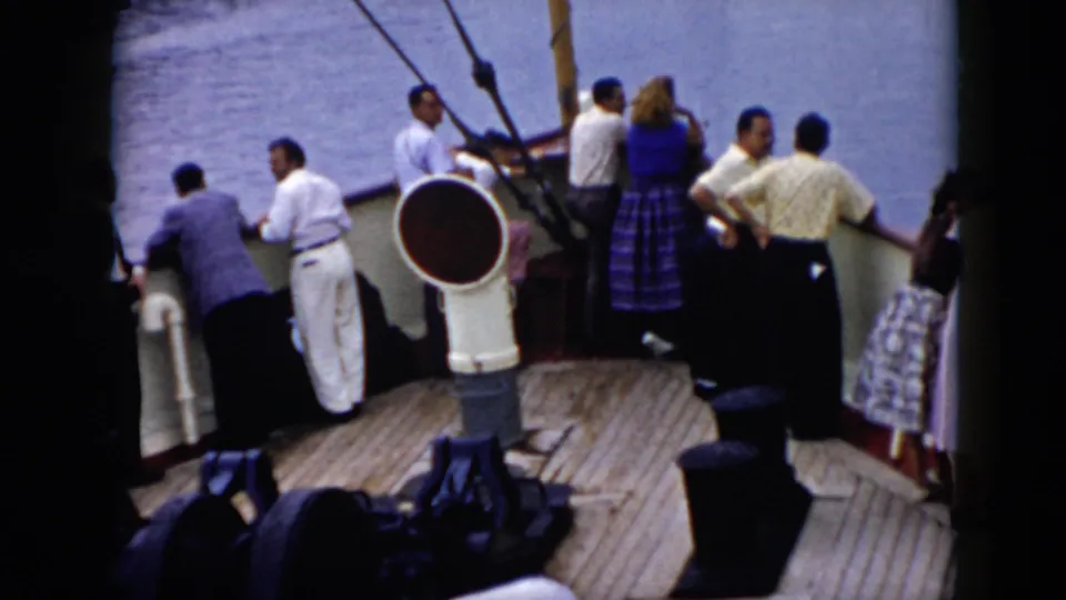 A cruise ship deck scene shows adults in light clothing and dresses standing near the rail with water beyond.